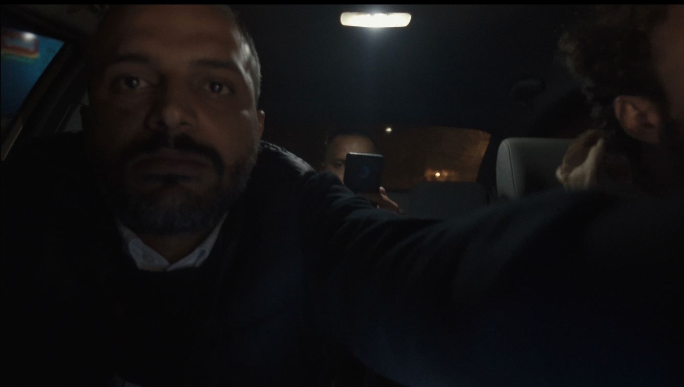 A man reaches toward the camera inside a dark car while someone behind him films with a phone.