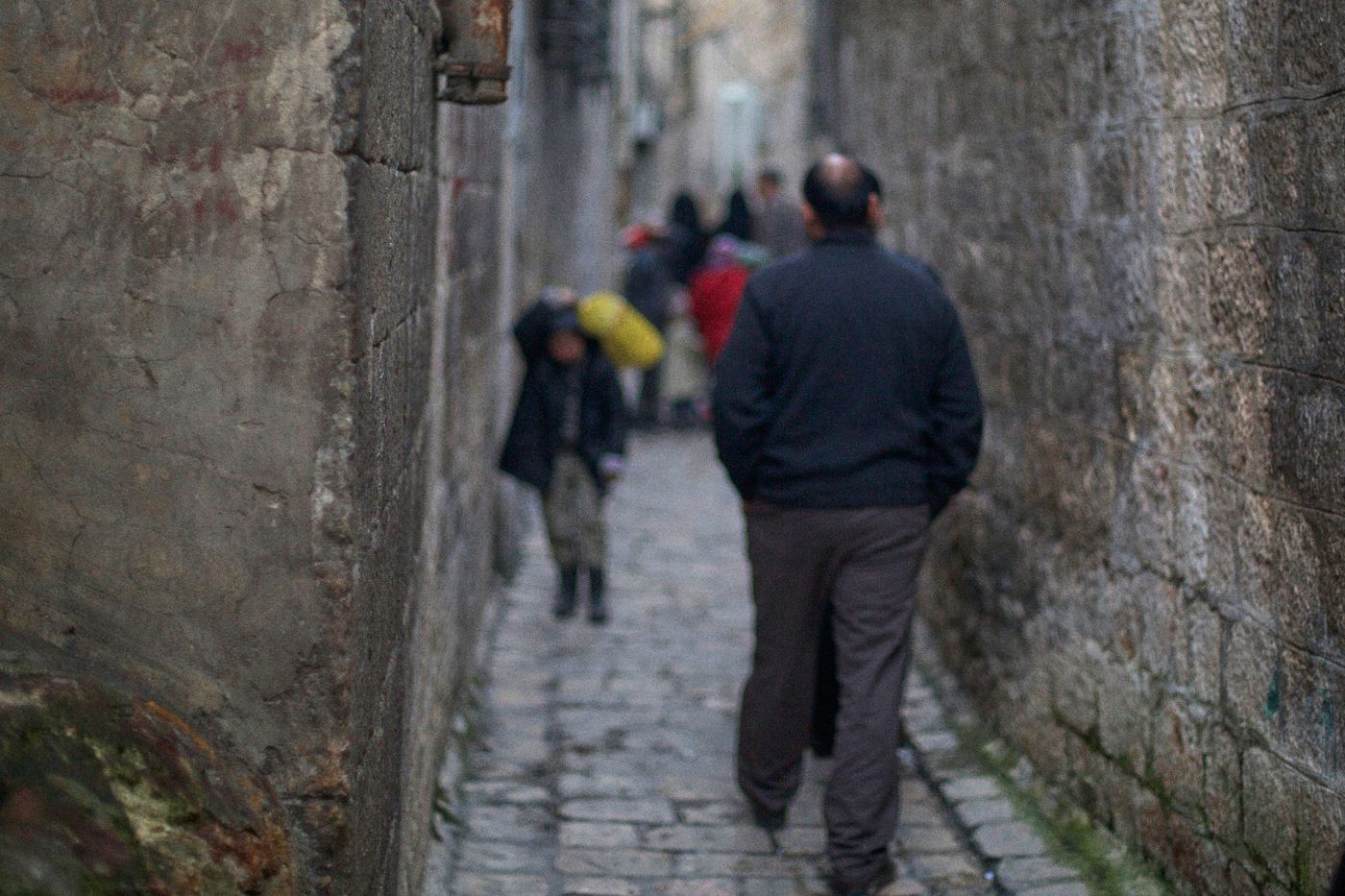 A man in a dark jacket walks away down a narrow stone alley, out of focus.