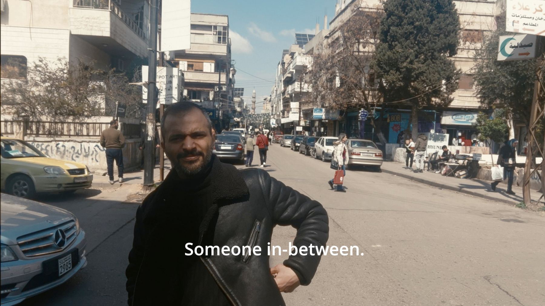 A man in a shearling jacket stands on a busy street, hand on hip. The subtitle reads: Someone in-between.