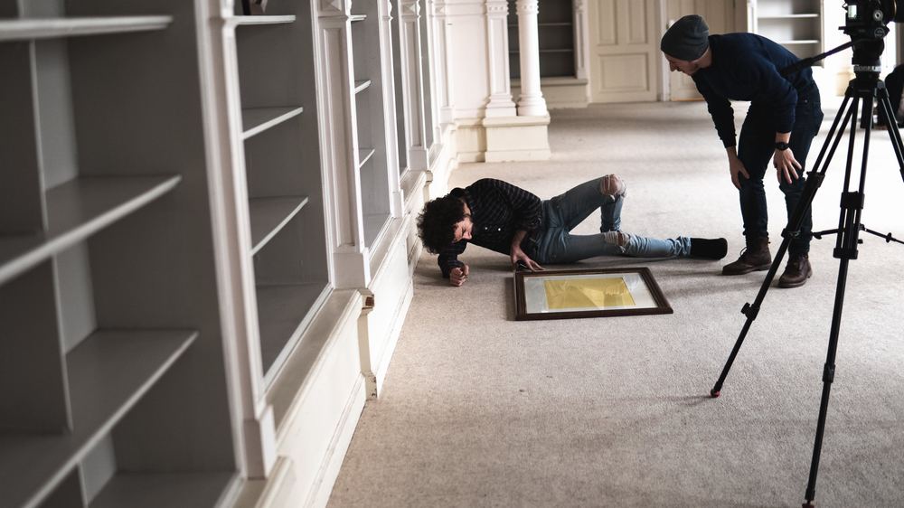 On a film set, a man lies on the carpet looking at a framed picture while another watches beside a tripod.