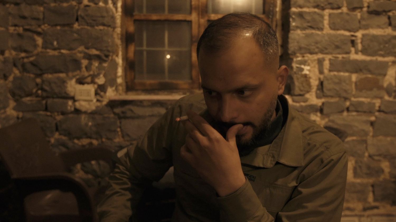 A man holds a cigarette near his face in a stone-walled café, eyes turned aside.