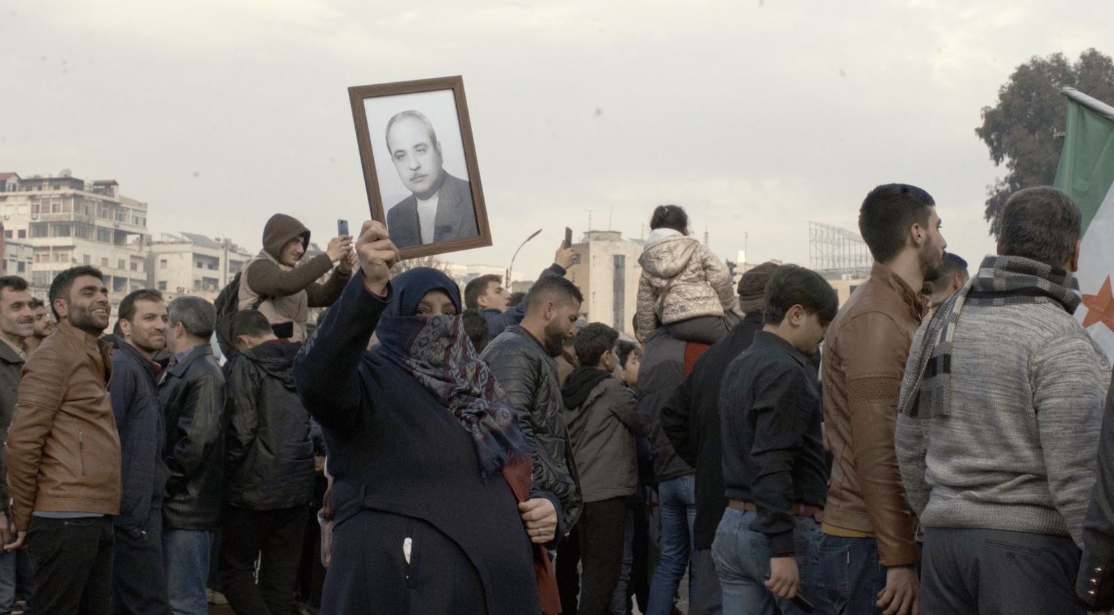 In a crowd, a veiled woman holds up a framed portrait of an older man.