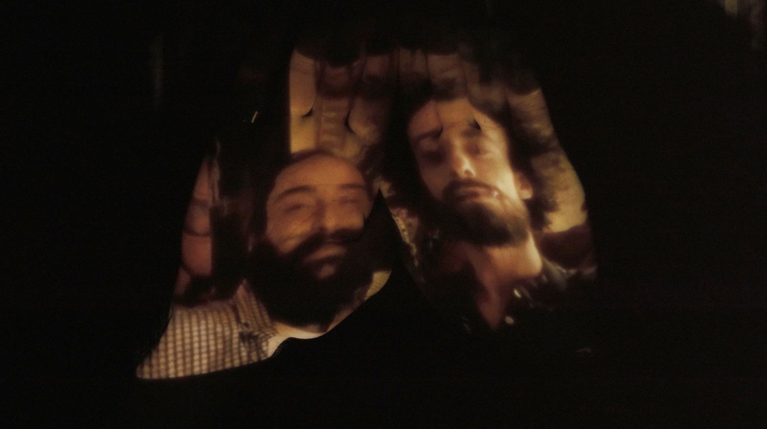Two bearded men’s faces glowing in warm light against black.
