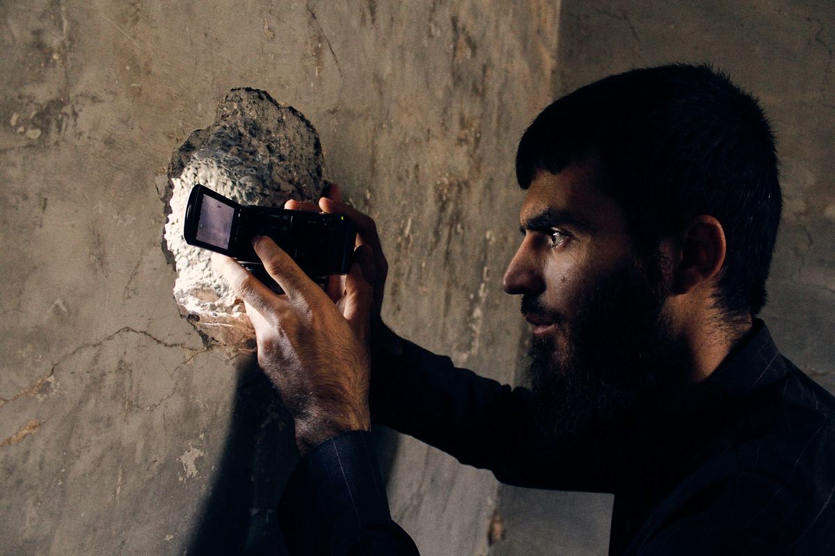 A bearded man holds a small camera up to a hole broken in a wall.