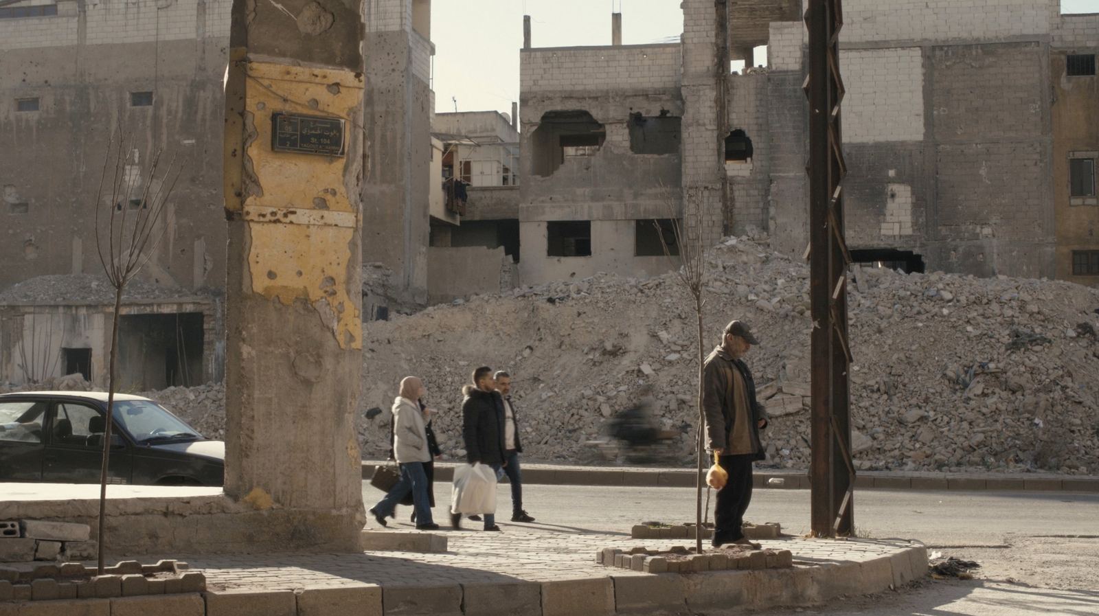 A man stands by a shell-scarred pillar in front of rubble and ruined buildings.