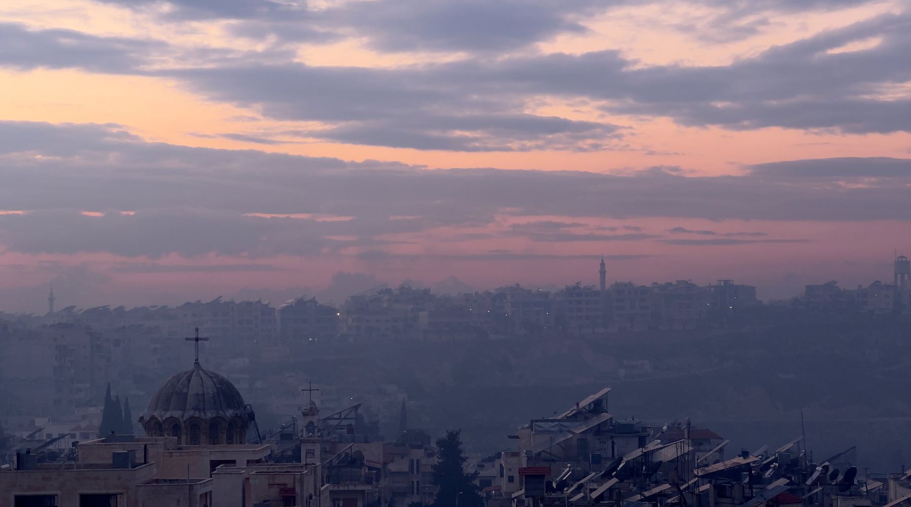 Dusk over a hillside city, pink sky, a church dome and rooftop solar panels in haze.
