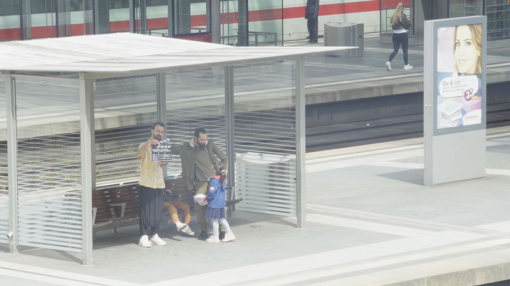 On a station platform, two men and a child under a shelter, one holding a clapperboard.