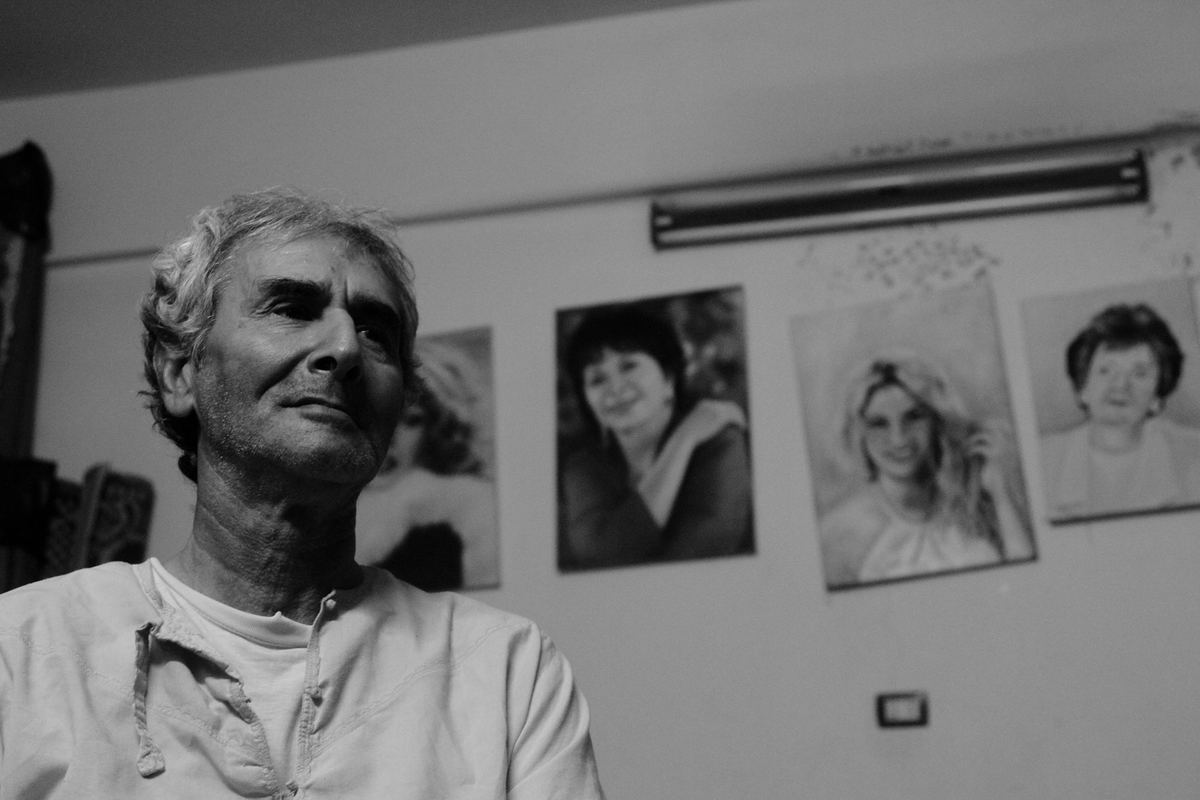 Black-and-white portrait of an older man looking away, with framed portraits of women on the wall behind him.
