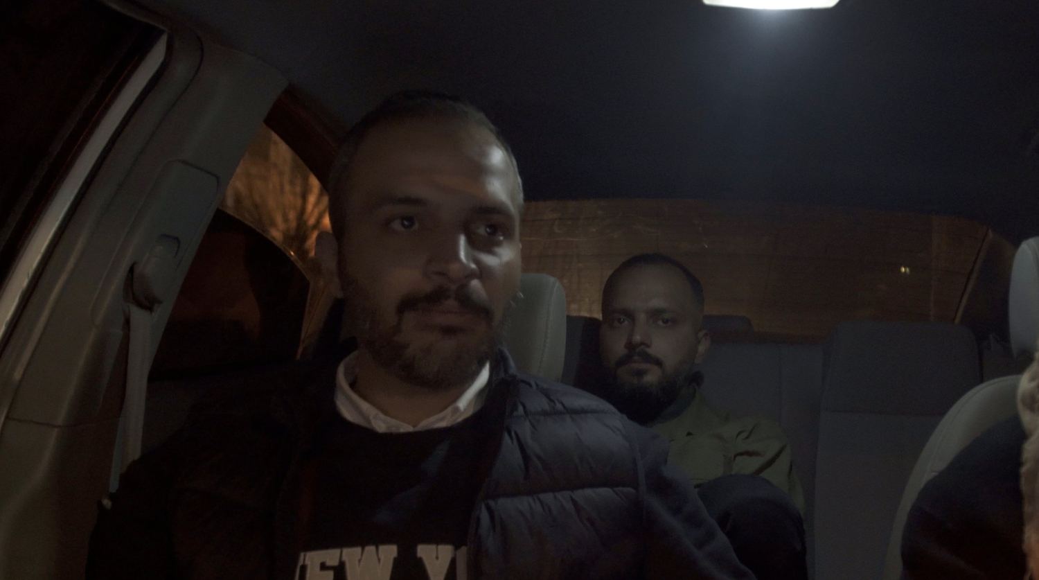 Two men in the front and back seats of a car at night, the dome light on.