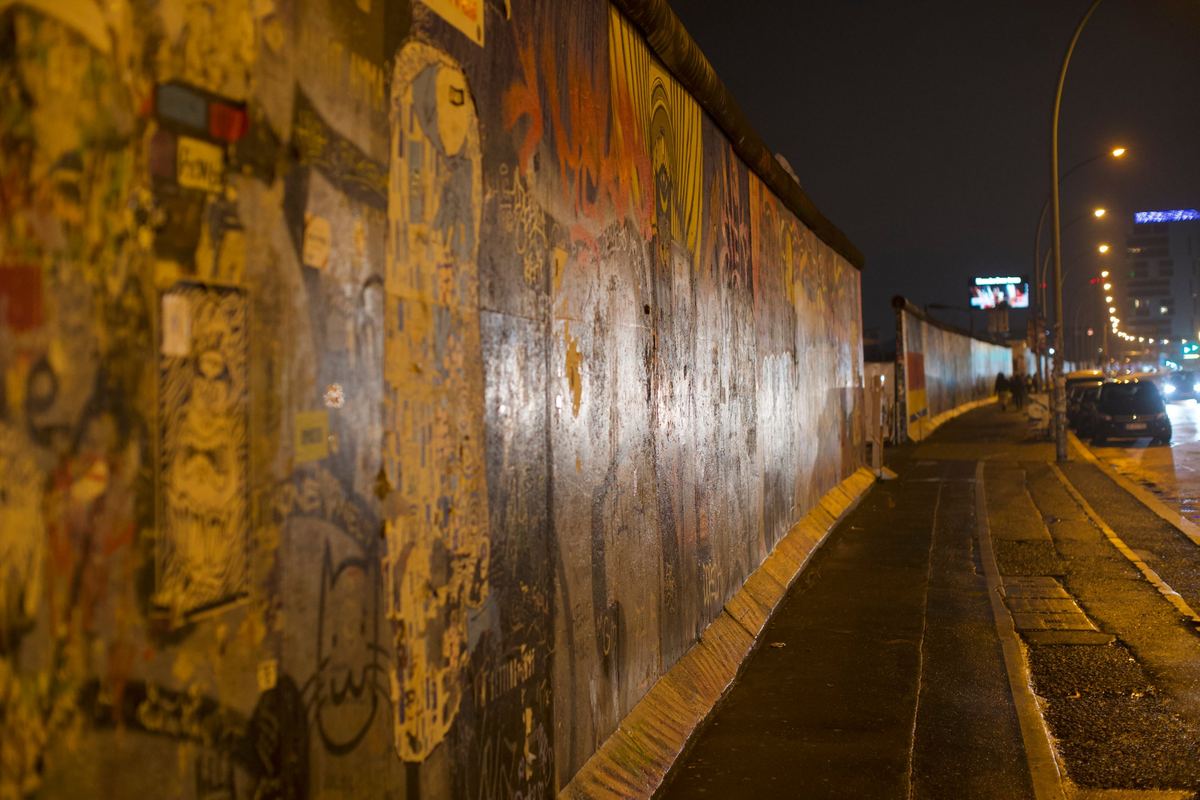 A long stretch of the Berlin Wall covered in graffiti, lit by street lamps at night.